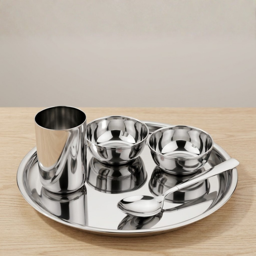 A stainless steel Indian-style plate set on a light wood table, featuring a round tray, a tall cup, two small bowls, and a spoon.